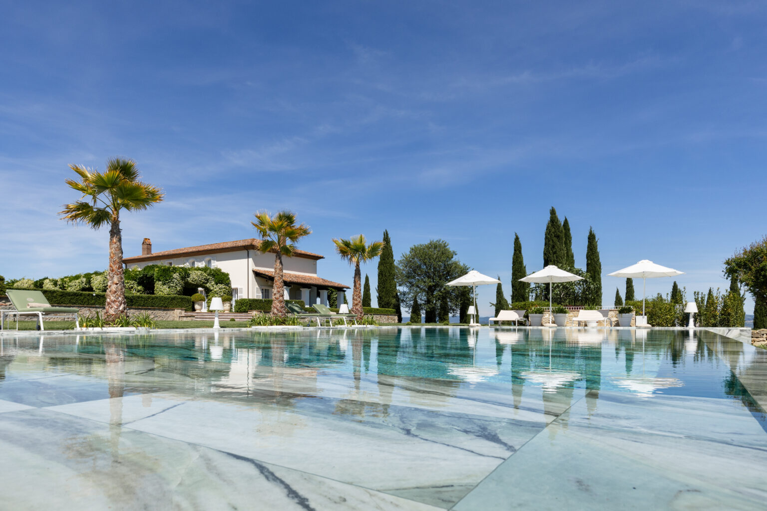 Luxurious villa with a swimming pool, featuring palm trees and sun loungers, surrounded by lush greenery and cypress trees under a clear blue sky. Ideal for relaxation and outdoor enjoyment. | Only on SopranoVillas