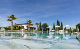 Luxurious villa with a swimming pool, featuring palm trees and sun loungers, surrounded by lush greenery and cypress trees under a clear blue sky. Ideal for relaxation and outdoor enjoyment.