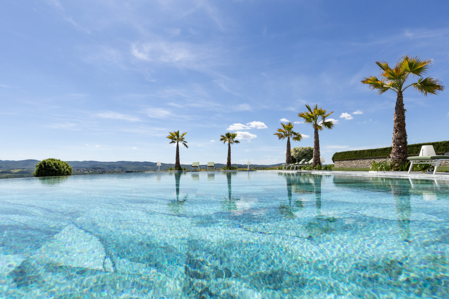 Infinity pool surrounded by palm trees, with a clear blue sky and distant mountains in the background, creating a serene and luxurious atmosphere. Ideal for relaxation and outdoor leisure activities. | Only on SopranoVillas