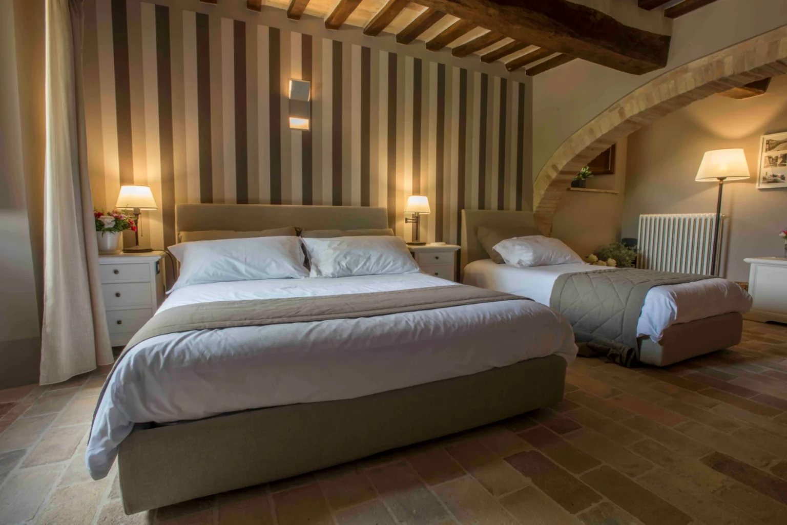 Cozy bedroom featuring a king-size bed and a twin bed, with soft linens and stylish bedding. The room is decorated with striped wallpaper and wooden beams, complemented by bedside lamps and a small nightstand, creating a warm and inviting atmosphere. | Only on SopranoVillas