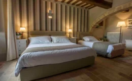 Cozy bedroom featuring a king-size bed and a twin bed, with soft linens and stylish bedding. The room is decorated with striped wallpaper and wooden beams, complemented by bedside lamps and a small nightstand, creating a warm and inviting atmosphere.
