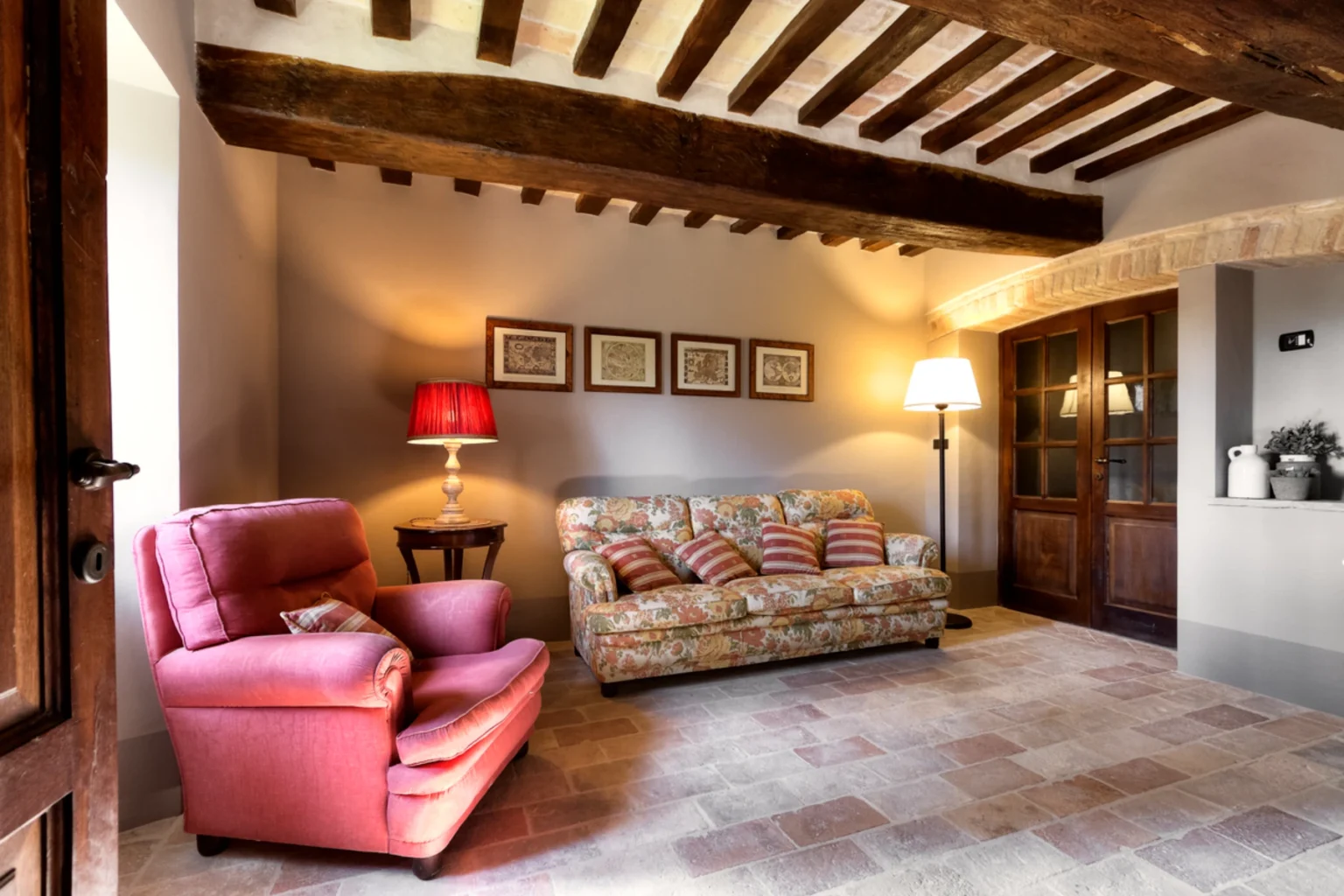 Cozy living room with a floral sofa and pink armchair, featuring wooden beams and warm lighting. The space includes decorative wall art, a side table with a lamp, and a view of a wooden door, highlighting a rustic yet inviting atmosphere. | Only on SopranoVillas