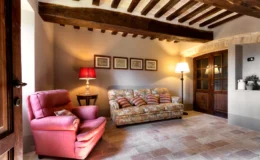 Cozy living room with a floral sofa and pink armchair, featuring wooden beams and warm lighting. The space includes decorative wall art, a side table with a lamp, and a view of a wooden door, highlighting a rustic yet inviting atmosphere.
