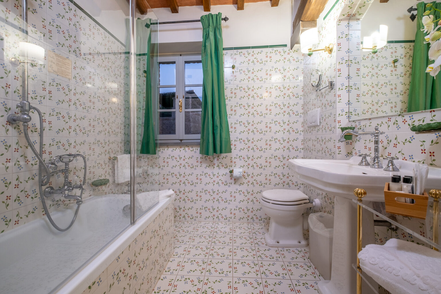 Bright and stylish bathroom featuring a bathtub with glass enclosure, floral patterned tiles, a white sink with elegant fixtures, and green curtains. The space is well-lit with natural light from a window, creating a welcoming atmosphere. | Only on SopranoVillas