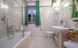 Bright and stylish bathroom featuring a bathtub with glass enclosure, floral patterned tiles, a white sink with elegant fixtures, and green curtains. The space is well-lit with natural light from a window, creating a welcoming atmosphere.