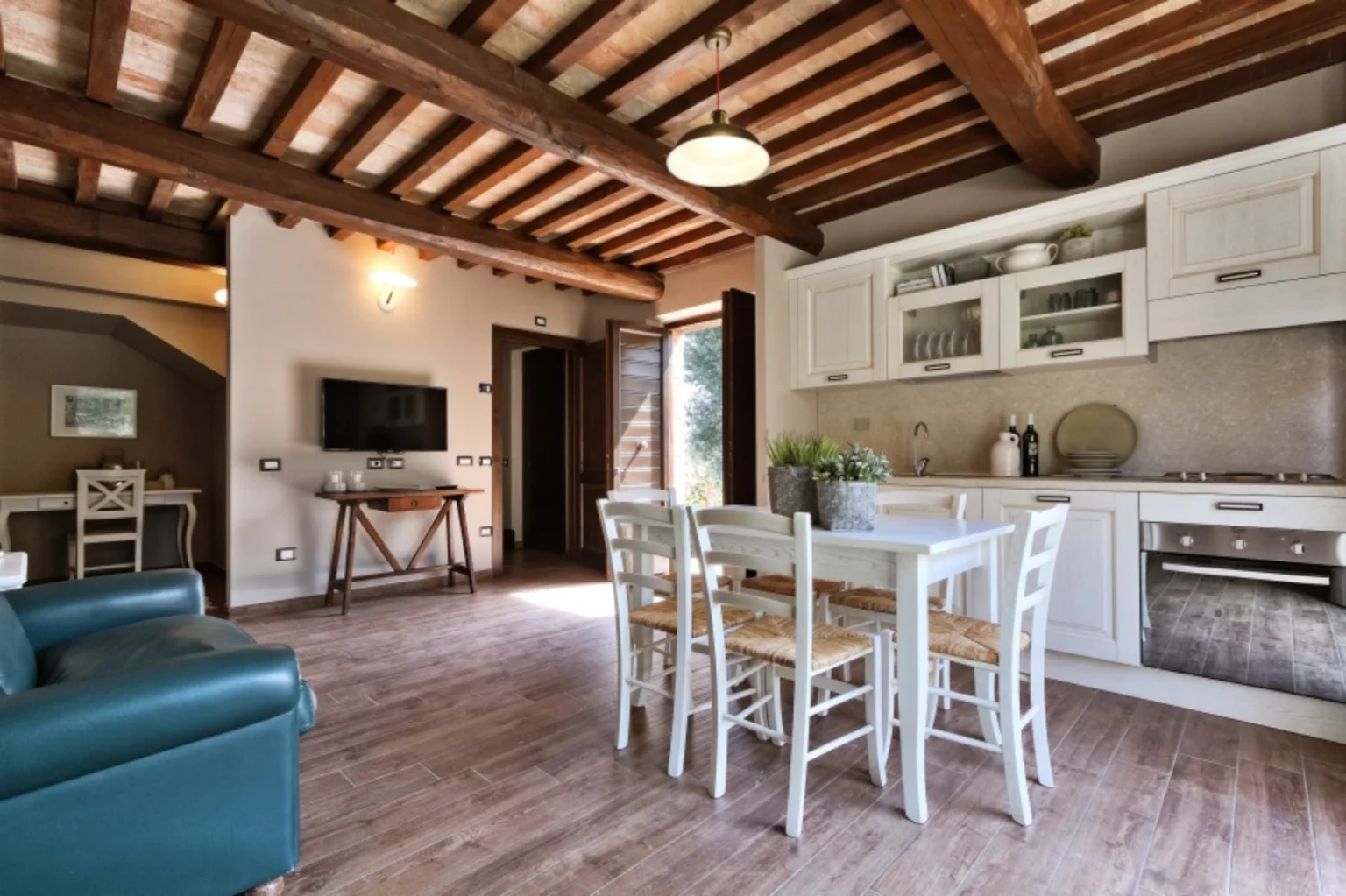 Modern living room and kitchen area featuring wooden beams, a dining table with chairs, a stylish kitchen with white cabinets, and a cozy seating arrangement. Natural light floods the space, enhancing the inviting atmosphere. | Only on SopranoVillas