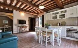 Modern living room and kitchen area featuring wooden beams, a dining table with chairs, a stylish kitchen with white cabinets, and a cozy seating arrangement. Natural light floods the space, enhancing the inviting atmosphere.