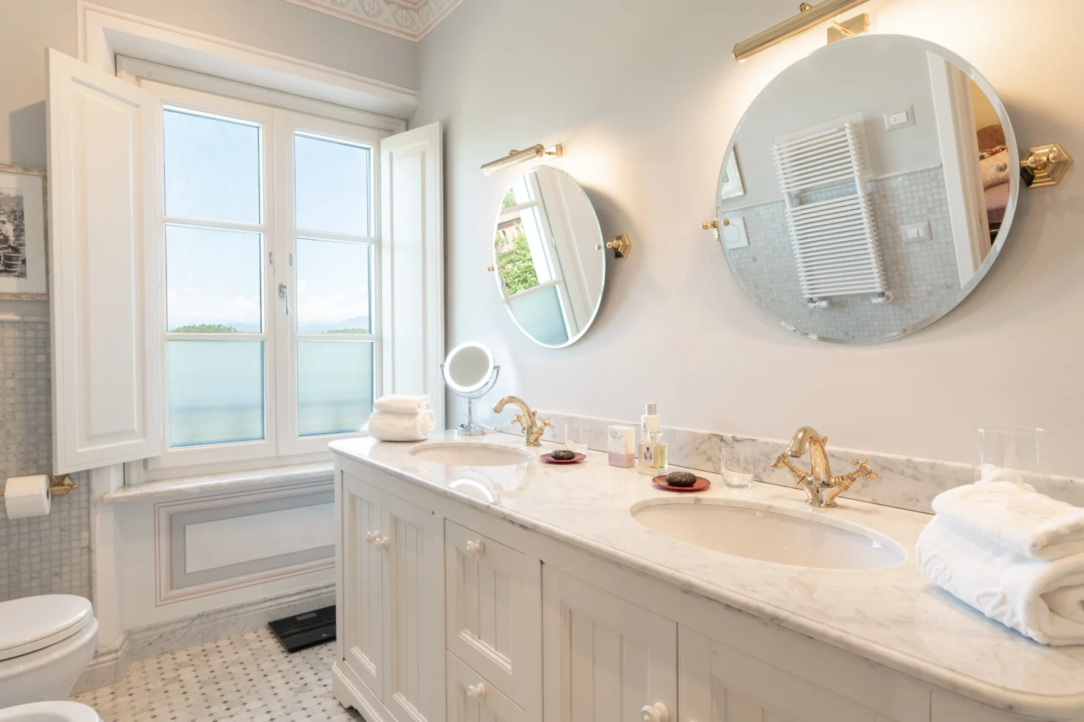 Luxury villa bathroom with elegant marble countertops, double sinks, and large mirrors, featuring a scenic view through the window, ideal for a relaxing stay in Italy. | Only on SopranoVillas