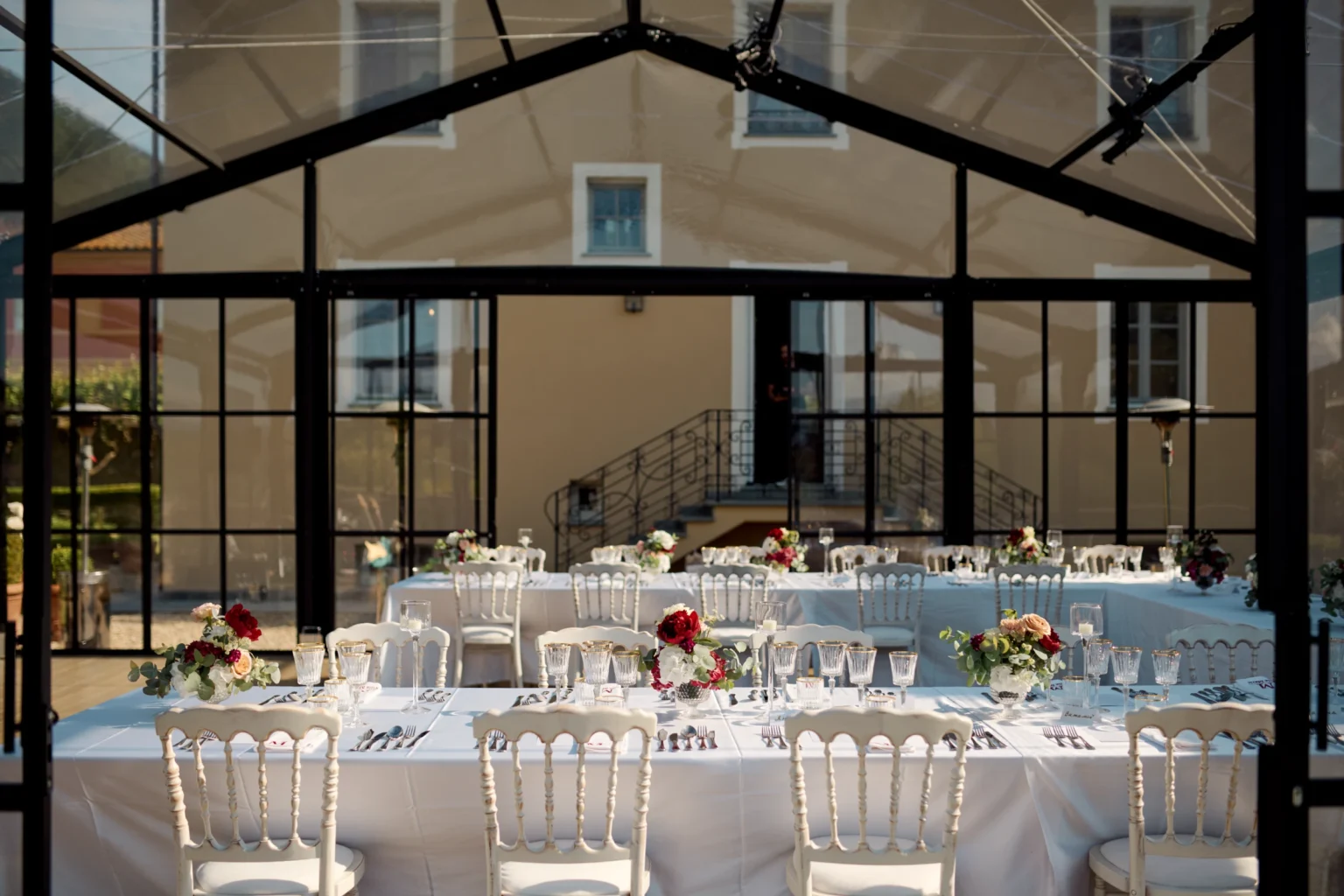Elegant dining setup in a luxury villa in Italy, featuring a beautifully arranged table with white tablecloth, fine dining utensils, and floral centerpieces, set in a stylish glass-enclosed space ideal for events and gatherings. | Only on SopranoVillas