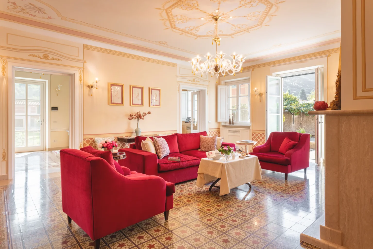 Elegant living room in a luxury villa for rent in Italy, featuring red upholstered furniture, a chandelier, and large windows that offer natural light and a view of the garden. Perfect for a sophisticated getaway. | Only on SopranoVillas