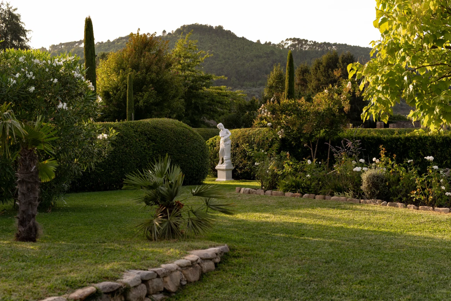 Lush garden view of a luxury villa in Italy featuring manicured lawns, ornamental trees, and a serene statue, perfect for a tranquil getaway. | Only on SopranoVillas