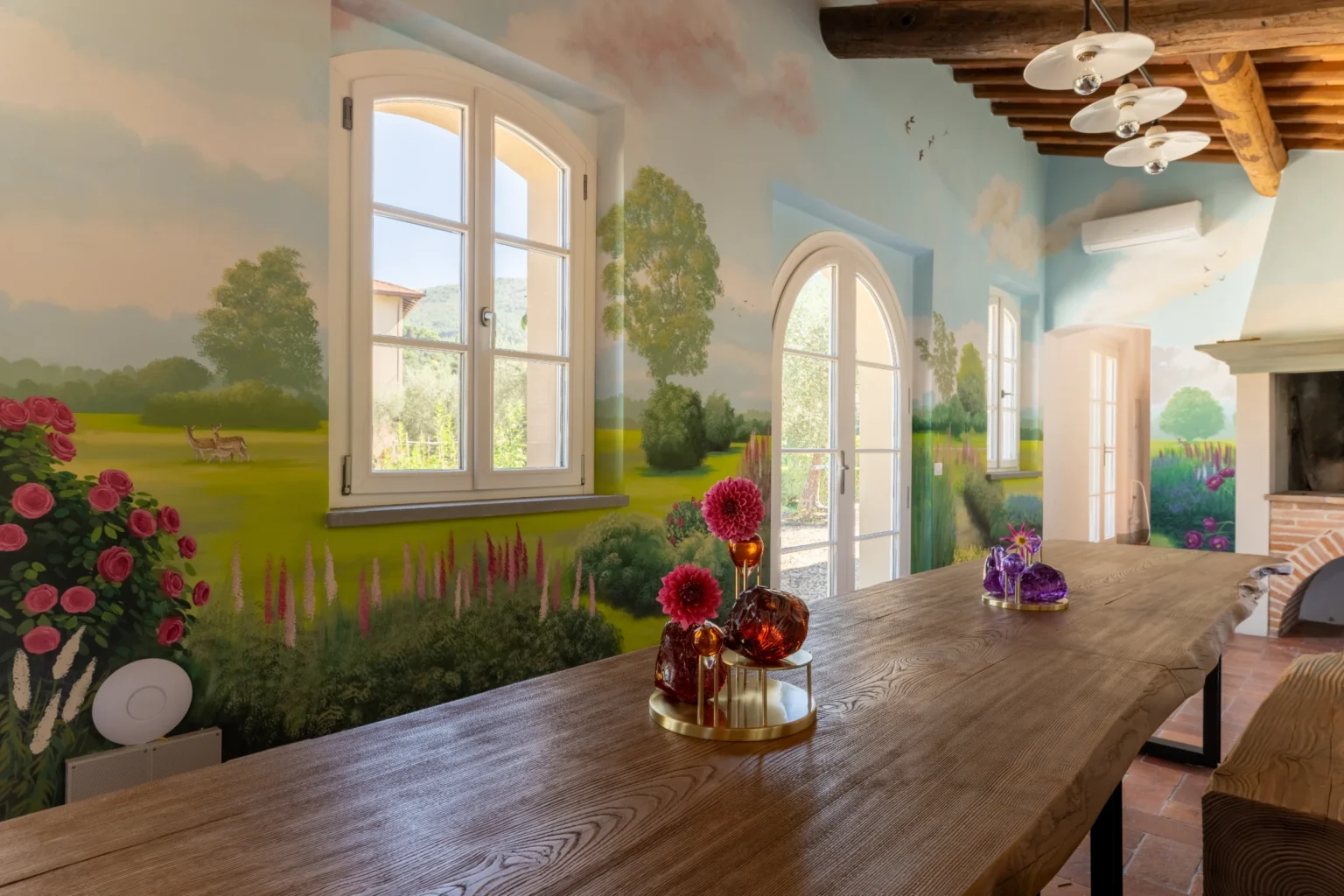Elegant dining area in a luxury villa for rent in Italy, featuring a beautifully painted mural, large windows with scenic views, and a rustic wooden table adorned with fresh flowers. | Only on SopranoVillas
