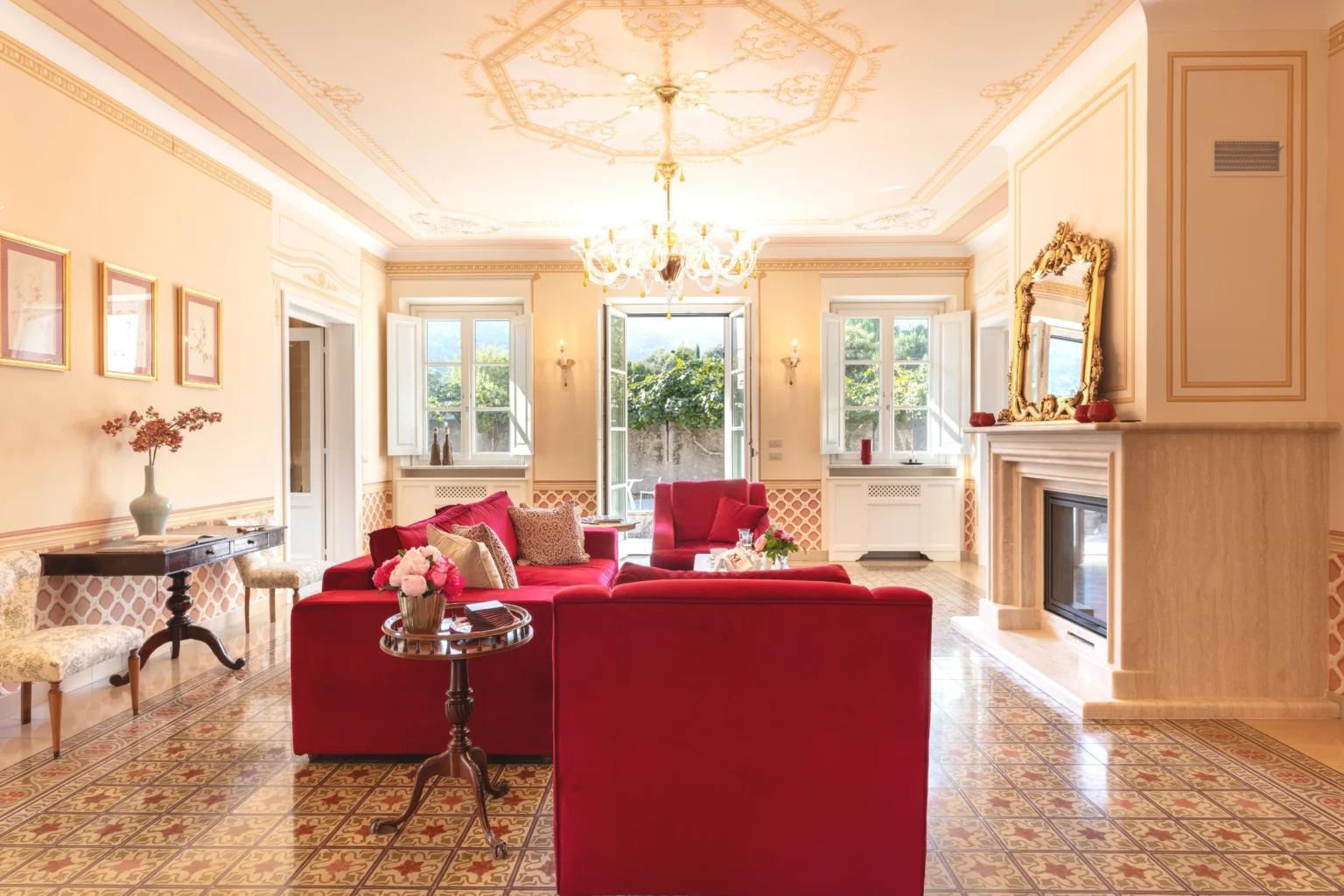 Luxurious interior of a villa in Italy featuring elegant red sofas, a chandelier, and large windows that open to a beautiful garden, perfect for vacation rentals. | Only on SopranoVillas