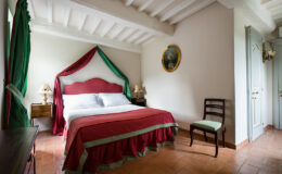 Cozy bedroom featuring a red and green bedspread, wooden furniture, and decorative curtains. The room has a classic design with exposed beams and terracotta flooring, creating an inviting atmosphere. A small bedside lamp and a framed painting add charm to the space.