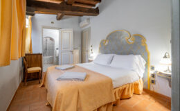 Cozy bedroom featuring a decorative yellow and gray headboard, white bedding, and an orange bedspread. The room includes wooden furniture, warm lighting, and a view of an adjoining bathroom with patterned tiles. Natural light filters in through yellow curtains, enhancing the inviting atmosphere. Ideal for a relaxing getaway.