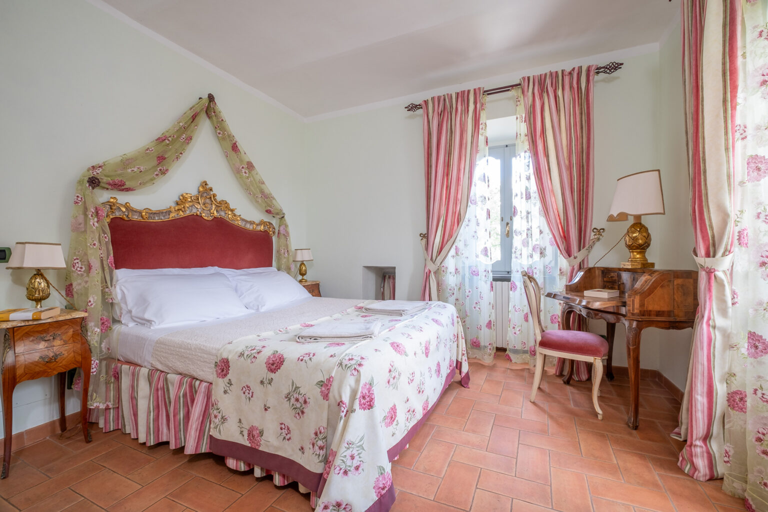 Elegant bedroom featuring a vintage design with a plush red headboard, floral-patterned bedding, and delicate drapes. The room includes a wooden desk with a pink chair and is adorned with soft lighting, creating a cozy atmosphere. Terracotta flooring complements the classic decor, making it an inviting space for relaxation. | Only on SopranoVillas