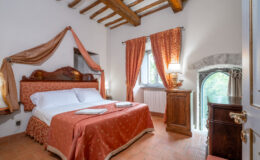 Cozy bedroom featuring a double bed with elegant bedding, wooden beams on the ceiling, and decorative curtains. The room includes a bedside lamp, a wooden nightstand, and a charming window with a view of greenery, creating a warm and inviting atmosphere.
