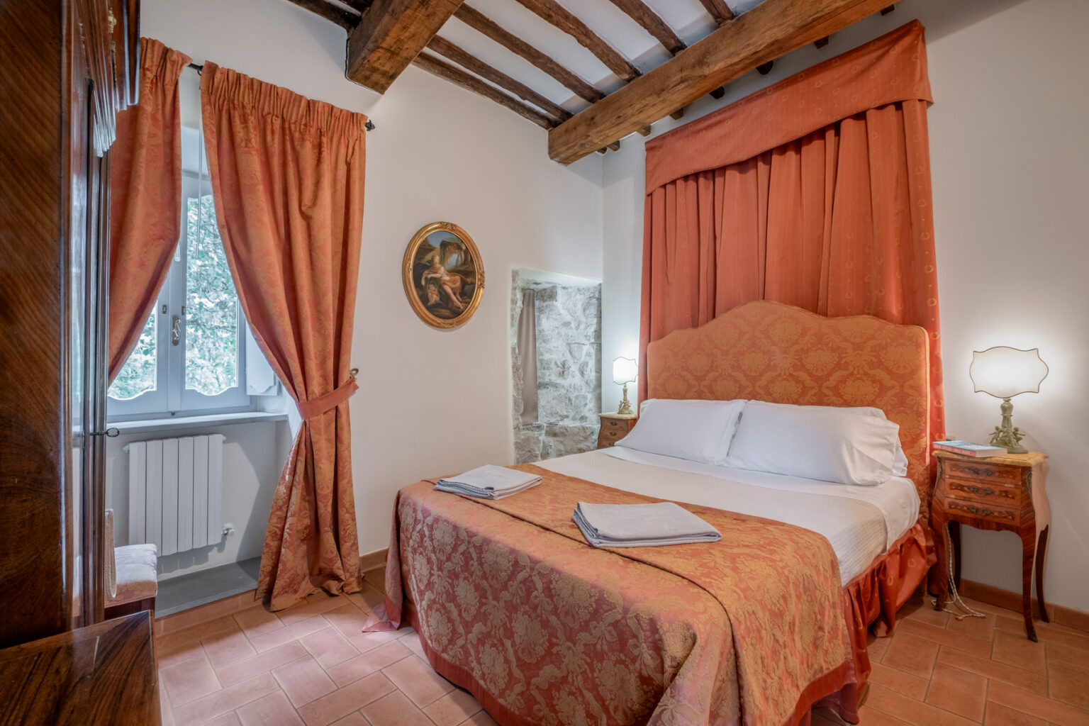 Cozy bedroom featuring a double bed with floral bedding, elegant curtains, and wooden beams. The room includes bedside lamps, a decorative painting, and a window with a view, creating a warm and inviting atmosphere. Ideal for a relaxing getaway. | Only on SopranoVillas