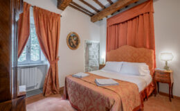 Cozy bedroom featuring a double bed with floral bedding, elegant curtains, and wooden beams. The room includes bedside lamps, a decorative painting, and a window with a view, creating a warm and inviting atmosphere. Ideal for a relaxing getaway.