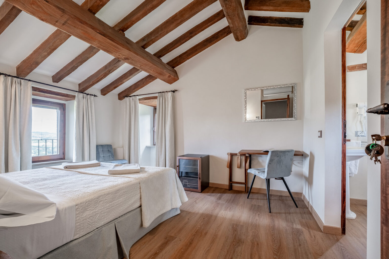Cozy bedroom featuring wooden beams, a comfortable bed with white linens, and natural light from two windows. Includes a desk and chair, a small cabinet, and a mirror, creating a serene and inviting atmosphere. | Only on SopranoVillas