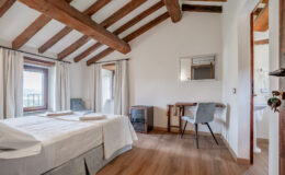 Cozy bedroom featuring wooden beams, a comfortable bed with white linens, and natural light from two windows. Includes a desk and chair, a small cabinet, and a mirror, creating a serene and inviting atmosphere.