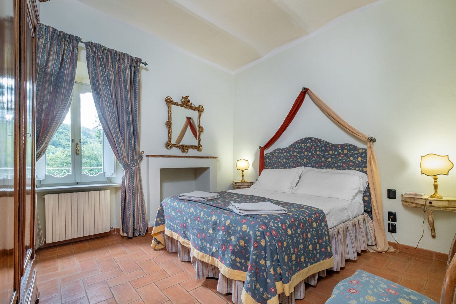 Cozy bedroom featuring a floral-patterned bedspread, elegant furnishings, and large windows with scenic views. The room includes a comfortable double bed, decorative curtains, a classic mirror, and ambient lighting, creating a warm and inviting atmosphere. Perfect for a relaxing stay. | Only on SopranoVillas