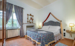 Cozy bedroom featuring a floral-patterned bedspread, elegant furnishings, and large windows with scenic views. The room includes a comfortable double bed, decorative curtains, a classic mirror, and ambient lighting, creating a warm and inviting atmosphere. Perfect for a relaxing stay.