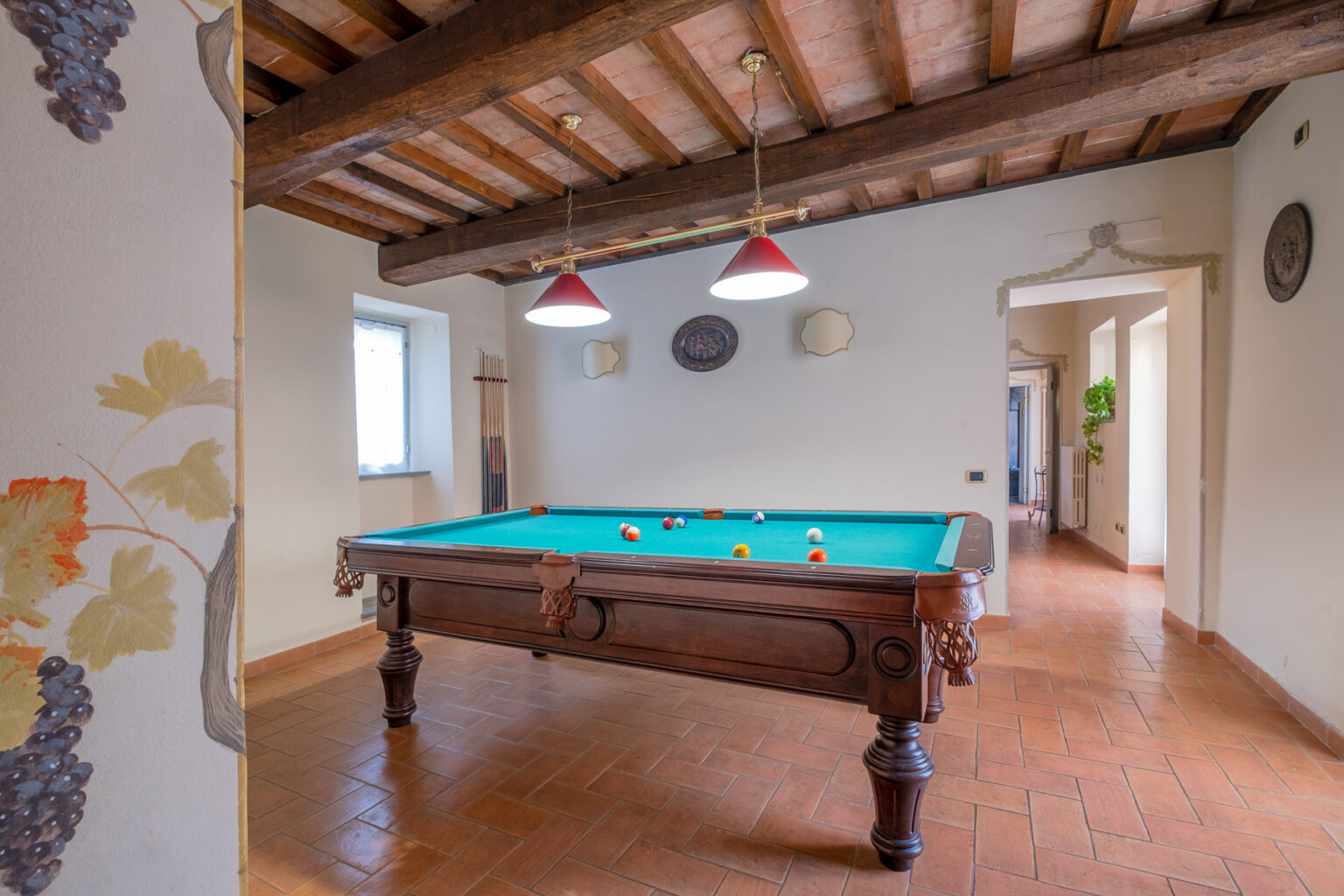 Interior view of a game room featuring a classic wooden billiards table with colorful balls, surrounded by terracotta tiled flooring and exposed wooden beams. Natural light enters through a window, enhancing the cozy atmosphere. | Only on SopranoVillas