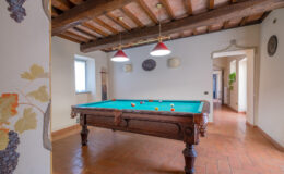 Interior view of a game room featuring a classic wooden billiards table with colorful balls, surrounded by terracotta tiled flooring and exposed wooden beams. Natural light enters through a window, enhancing the cozy atmosphere.