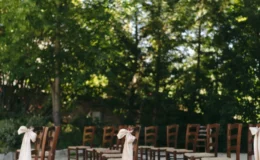 Outdoor wedding ceremony setup featuring wooden chairs adorned with elegant cream ribbons, surrounded by lush greenery and sunlight filtering through the trees. Ideal for a romantic and intimate wedding atmosphere.
