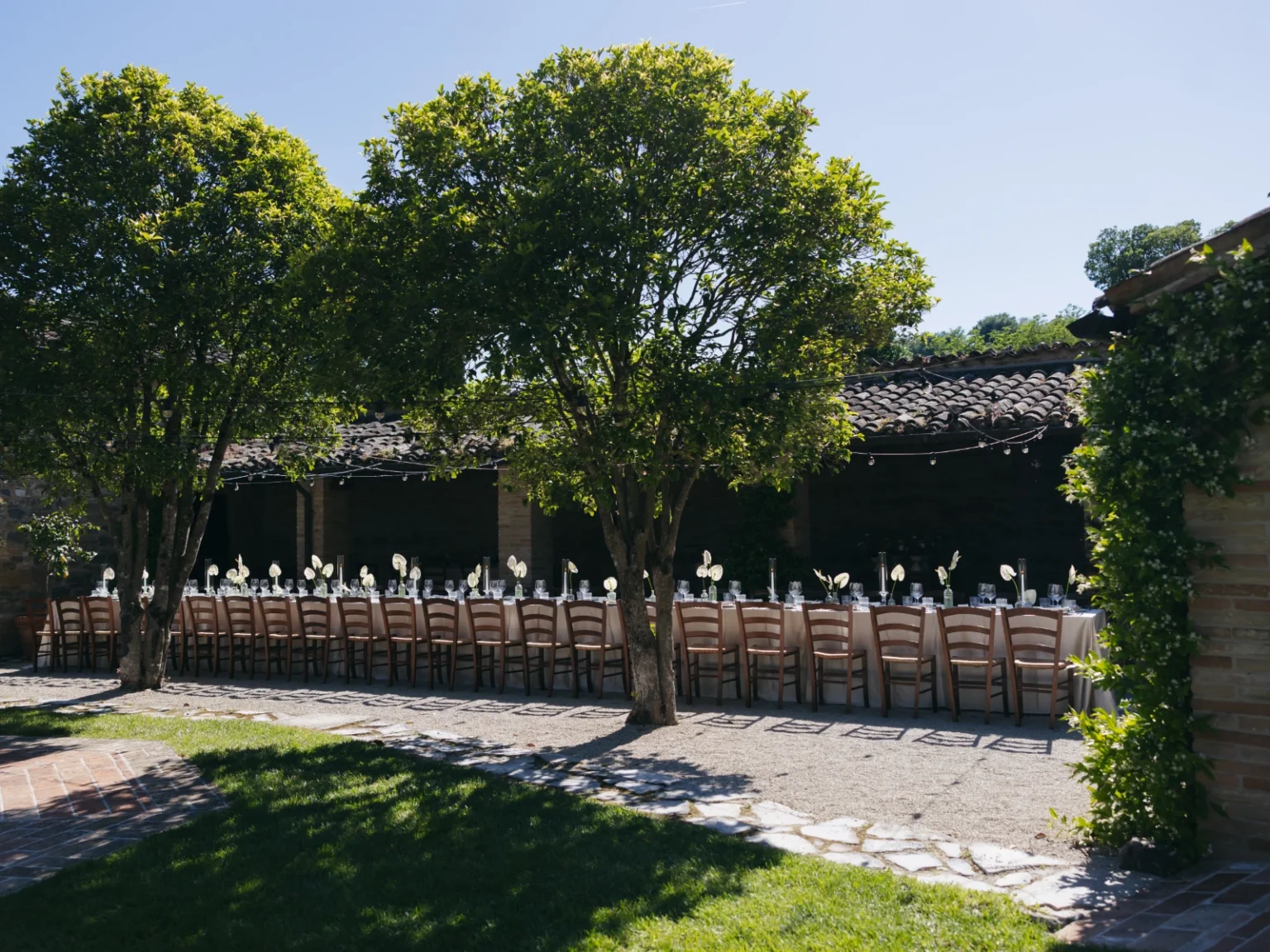 Outdoor dining setup under trees with elegant table settings, featuring white floral arrangements and glassware, in a scenic garden venue. Ideal for weddings and special events. | Only on SopranoVillas