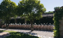 Outdoor dining setup under trees with elegant table settings, featuring white floral arrangements and glassware, in a scenic garden venue. Ideal for weddings and special events.