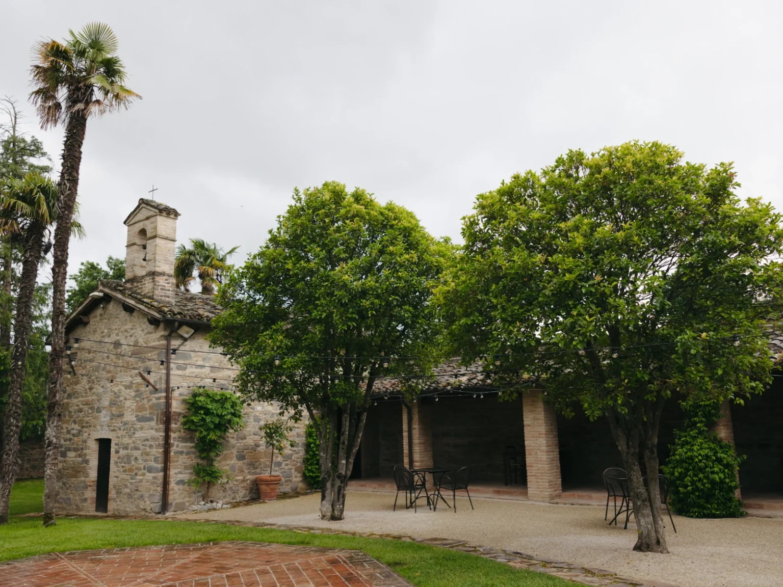 Stone building with a bell tower surrounded by lush greenery and palm trees, featuring outdoor seating and a charming garden area under a cloudy sky. | Only on SopranoVillas
