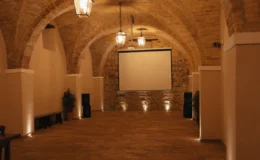 Interior of a rustic event space featuring arched brick ceilings, hanging lantern lights, and a large projection screen. The warm-toned tiled floor complements the elegant atmosphere, suitable for gatherings and presentations.