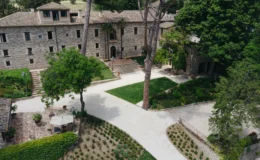 Aerial view of a charming stone villa surrounded by lush greenery, featuring a well-maintained garden, pathways, and outdoor seating areas. The villa's rustic architecture blends harmoniously with the natural landscape, creating a serene atmosphere ideal for relaxation and gatherings.
