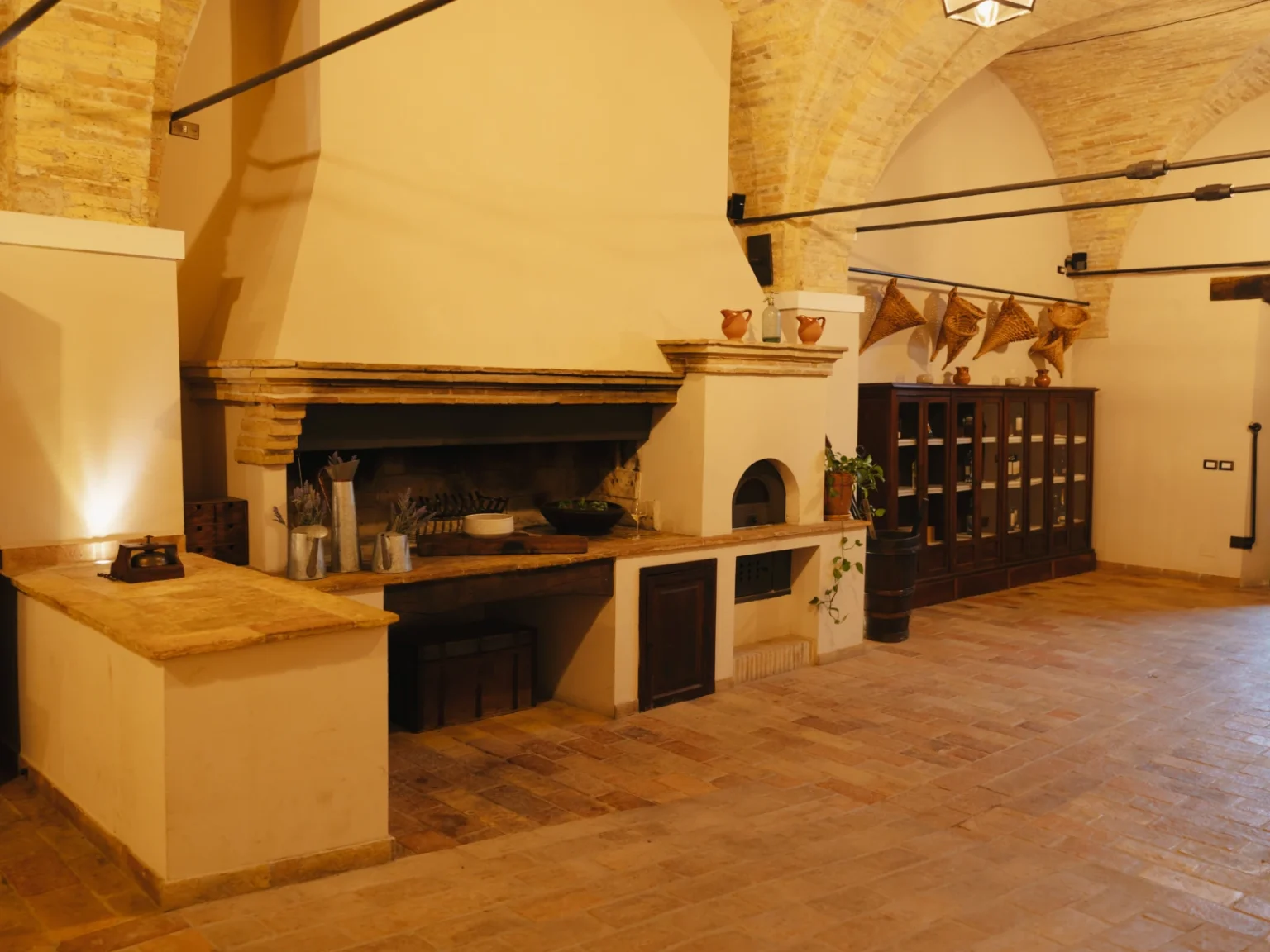 Rustic kitchen interior featuring a large stone fireplace, wooden cabinetry, and decorative elements like pottery and herbs. The space showcases arched ceilings and terracotta flooring, creating a warm and inviting atmosphere. Ideal for culinary gatherings and home cooking. | Only on SopranoVillas