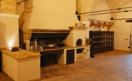 Rustic kitchen interior featuring a large stone fireplace, wooden cabinetry, and decorative elements like pottery and herbs. The space showcases arched ceilings and terracotta flooring, creating a warm and inviting atmosphere. Ideal for culinary gatherings and home cooking.