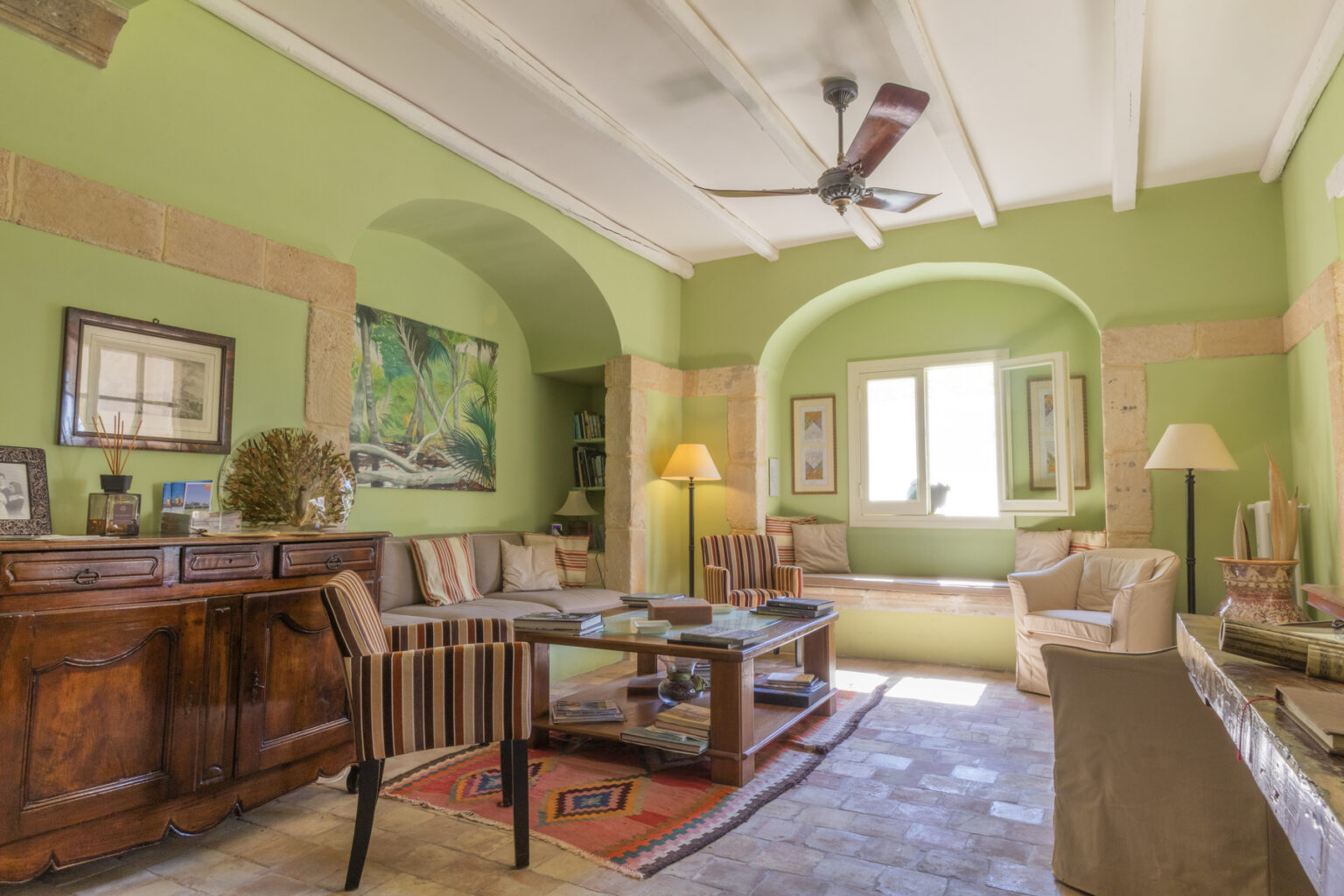 Bright and inviting living room featuring green walls, wooden furniture, and a ceiling fan. The space includes a comfortable seating area with striped chairs, a coffee table, and natural light streaming in through large windows. Decorative artwork and a cozy rug enhance the warm ambiance. | Only on SopranoVillas