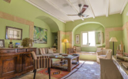 Bright and inviting living room featuring green walls, wooden furniture, and a ceiling fan. The space includes a comfortable seating area with striped chairs, a coffee table, and natural light streaming in through large windows. Decorative artwork and a cozy rug enhance the warm ambiance.