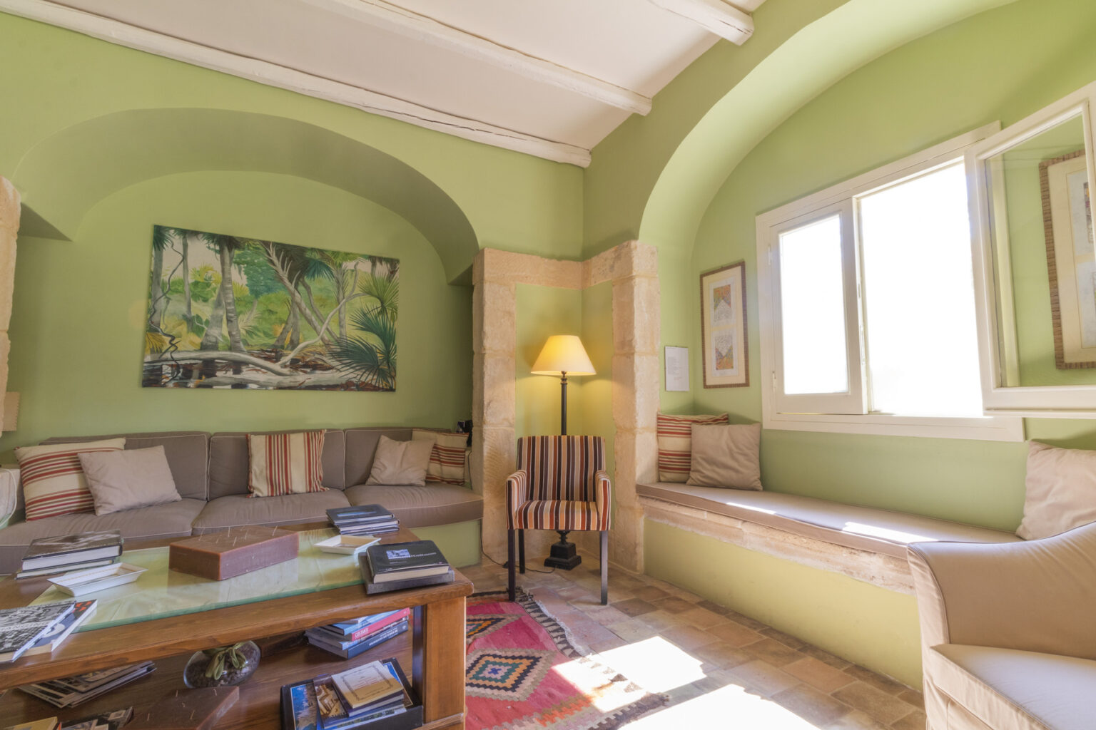Cozy living room featuring green walls, natural light from large windows, a comfortable sofa with decorative pillows, a wooden coffee table stacked with books, and an artistic wall painting. | Only on SopranoVillas