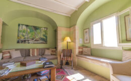 Cozy living room featuring green walls, natural light from large windows, a comfortable sofa with decorative pillows, a wooden coffee table stacked with books, and an artistic wall painting.