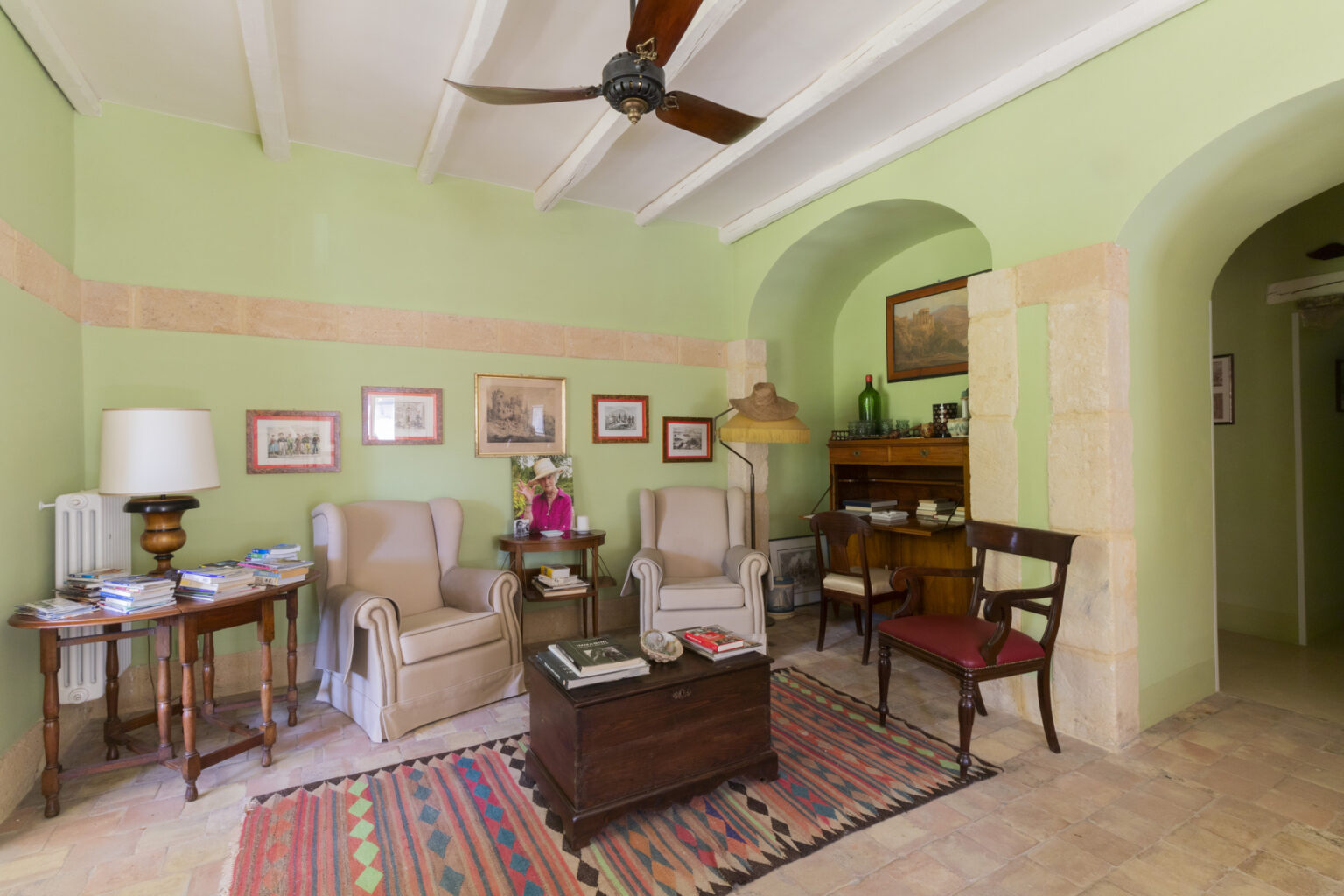 Cozy living room with green walls, featuring comfortable armchairs, a wooden coffee table, and decorative artwork. A ceiling fan and warm lighting create an inviting atmosphere, complemented by a colorful area rug. | Only on SopranoVillas