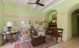 Cozy living room with green walls, featuring comfortable armchairs, a wooden coffee table, and decorative artwork. A ceiling fan and warm lighting create an inviting atmosphere, complemented by a colorful area rug.