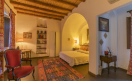 Cozy bedroom interior featuring a queen-sized bed, wooden ceiling beams, a vintage armchair, and decorative rugs, creating a warm and inviting atmosphere.