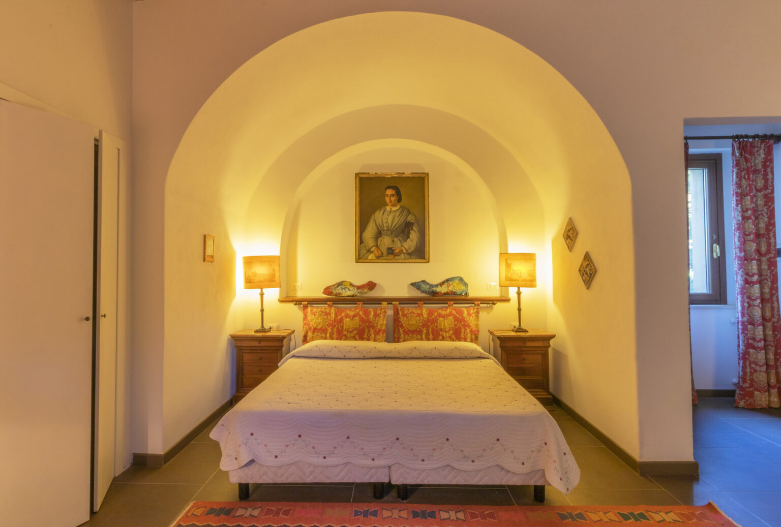 Cozy bedroom featuring a double bed with decorative pillows, warm lighting from bedside lamps, and an arched ceiling. The room includes a vintage painting and a window with patterned curtains, creating a welcoming atmosphere. | Only on SopranoVillas