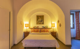 Cozy bedroom featuring a double bed with decorative pillows, warm lighting from bedside lamps, and an arched ceiling. The room includes a vintage painting and a window with patterned curtains, creating a welcoming atmosphere.