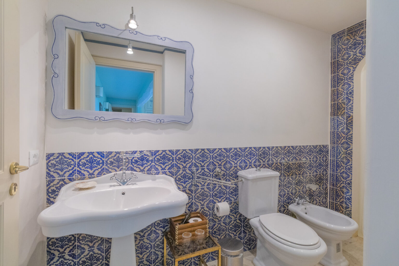 Modern bathroom featuring a stylish sink, bidet, and toilet against a blue and white patterned tiled wall, complemented by a decorative mirror and contemporary lighting. | Only on SopranoVillas