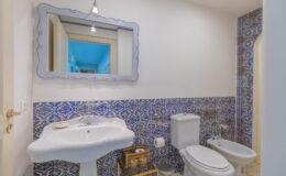 Modern bathroom featuring a stylish sink, bidet, and toilet against a blue and white patterned tiled wall, complemented by a decorative mirror and contemporary lighting.