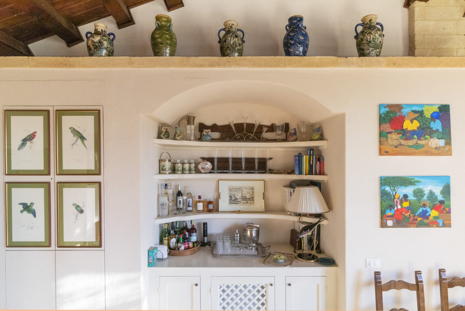 Interior view of a cozy living space featuring a decorative shelf with framed bird illustrations, a well-stocked bar area with various bottles, and colorful artwork depicting market scenes on the walls, all set against a warm, rustic backdrop. | Only on SopranoVillas