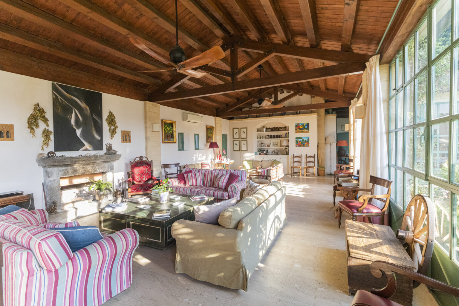 Bright and airy living room featuring wooden beams and large windows, furnished with striped sofas and a vintage red chair. Decorated with artwork, plants, and a stone fireplace, the space exudes a cozy and eclectic atmosphere. | Only on SopranoVillas